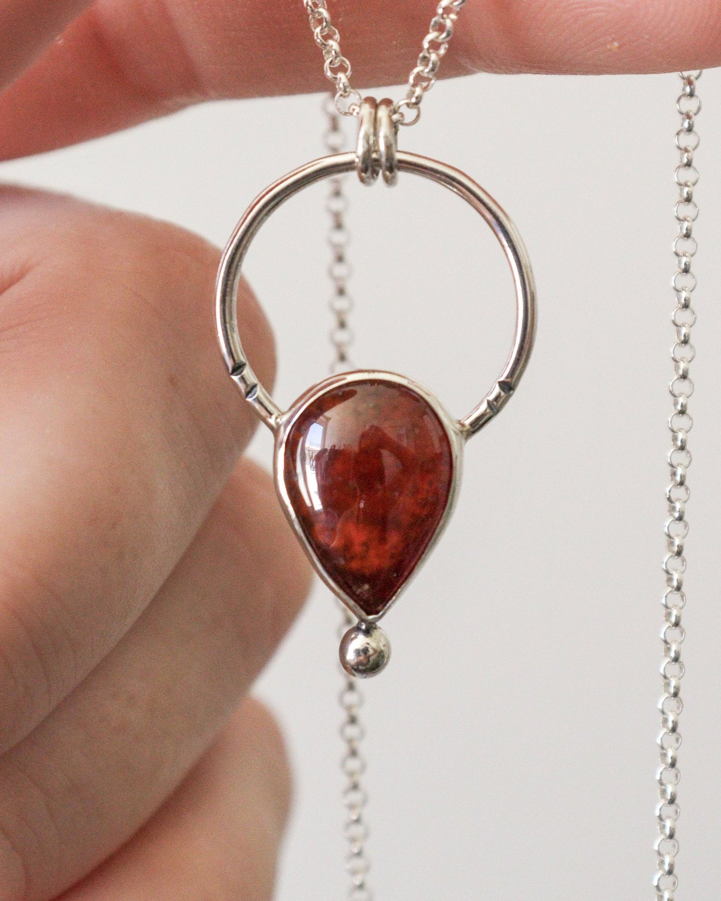 Un primer plano del colgante ARCO de plata con una piedra preciosa de granate rojo intenso en forma de lágrima, con una cadena de plata fina desenfocada en el fondo.