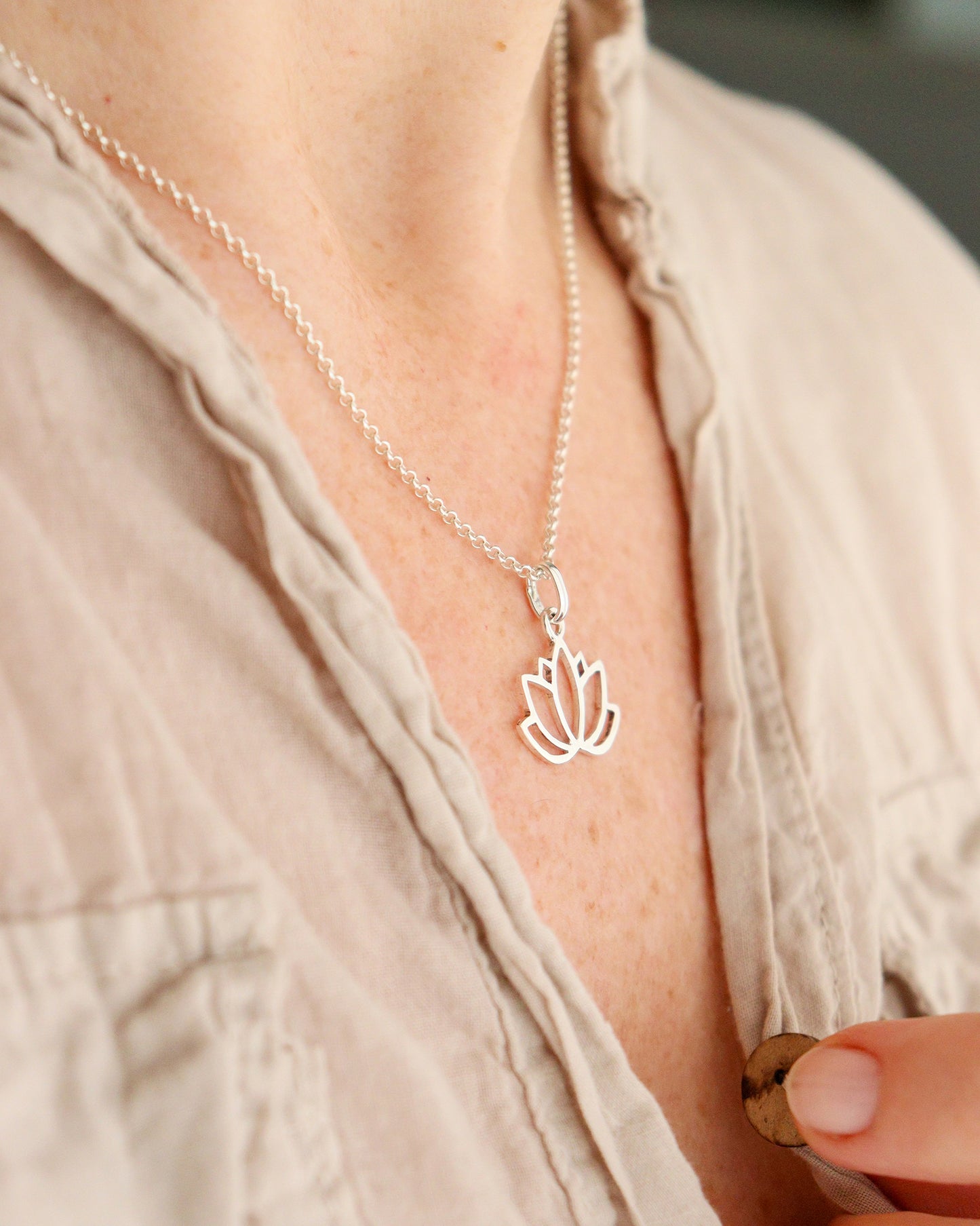 Una persona sostiene un collar con un colgante de plata en forma de flor de loto calada, mientras una parte de su cuello y hombro es visible con una camisa clara.