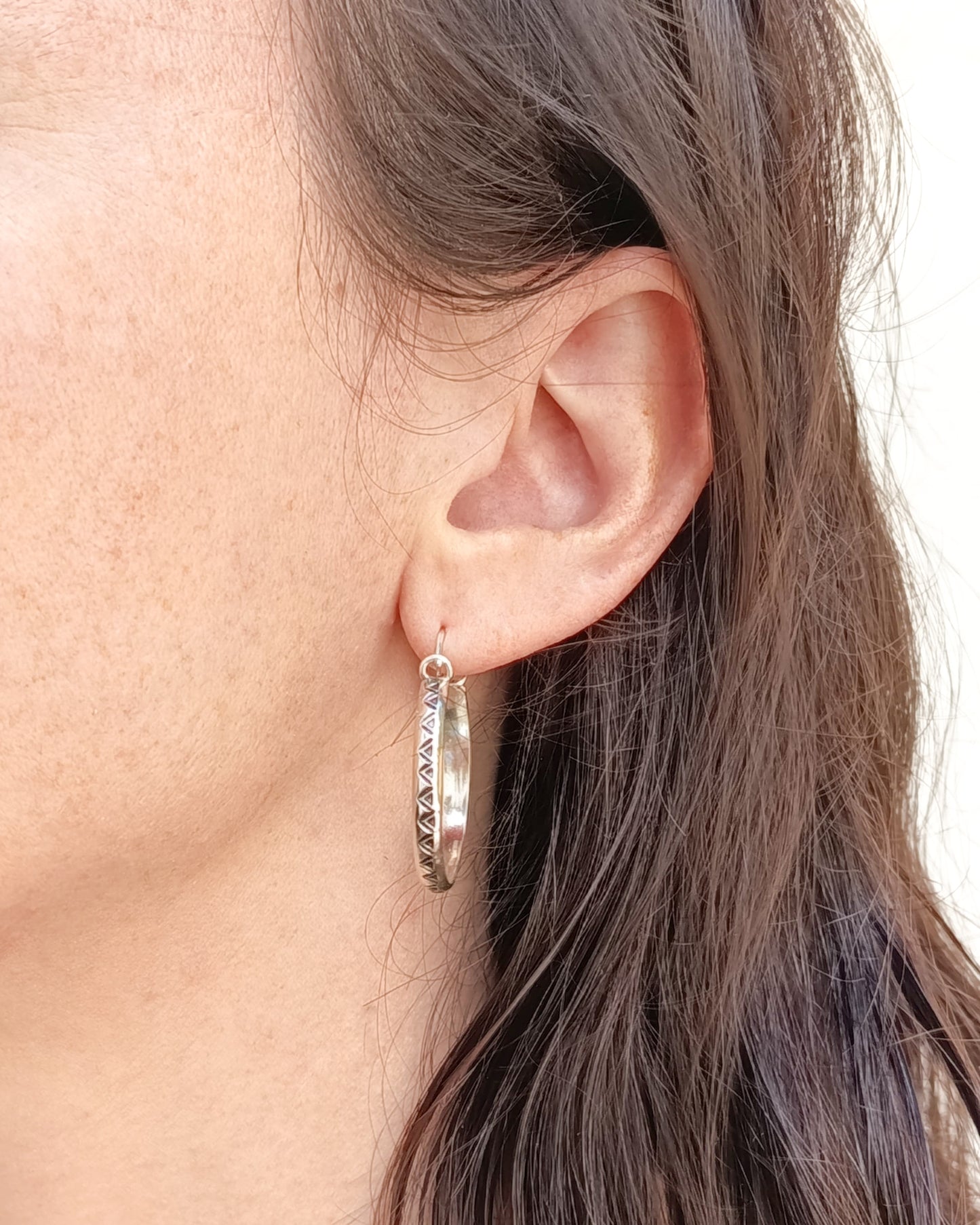 Primer plano de una oreja con un pendiente de aro plateado colgando del lóbulo. El pendiente tiene un patrón geométrico de triángulos grabado en su superficie (pendientes dde aro TRILO). Parte del cabello oscuro de la persona es visible al lado de la oreja. El fondo es claro.