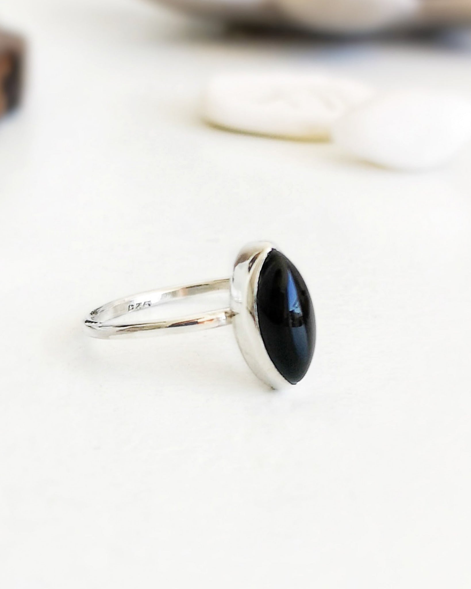 Anillo Esencial Mirar de plata con una piedra negra de forma marquise en el centro, visto de perfil sobre una superficie blanca con algunos objetos decorativos difuminados al fondo.