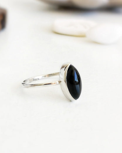 Anillo Esencial Mirar de plata con una piedra negra de forma marquise en el centro, visto de perfil sobre una superficie blanca con algunos objetos decorativos difuminados al fondo.
