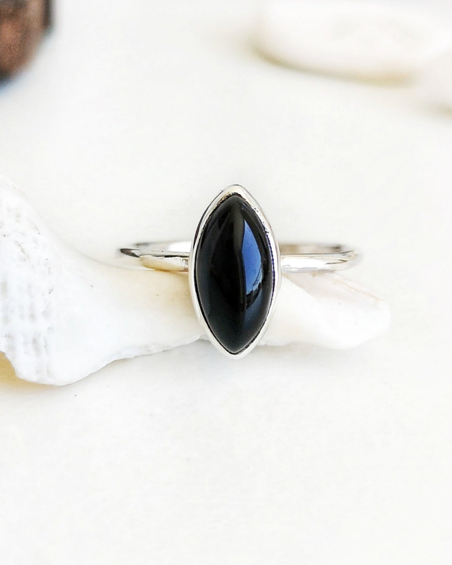 Anillo Esencial Mirar de plata con una piedra negra de forma marquiese en el centro, apoyado sobre una concha marina blanca con objetos decorativos de madera y otras conchas difuminados al fondo.