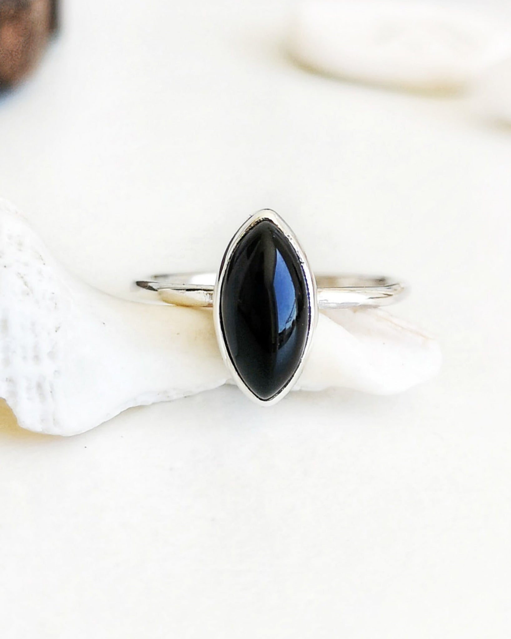 Anillo Esencial Mirar de plata con una piedra negra de forma marquiese en el centro, apoyado sobre una concha marina blanca con objetos decorativos de madera y otras conchas difuminados al fondo.