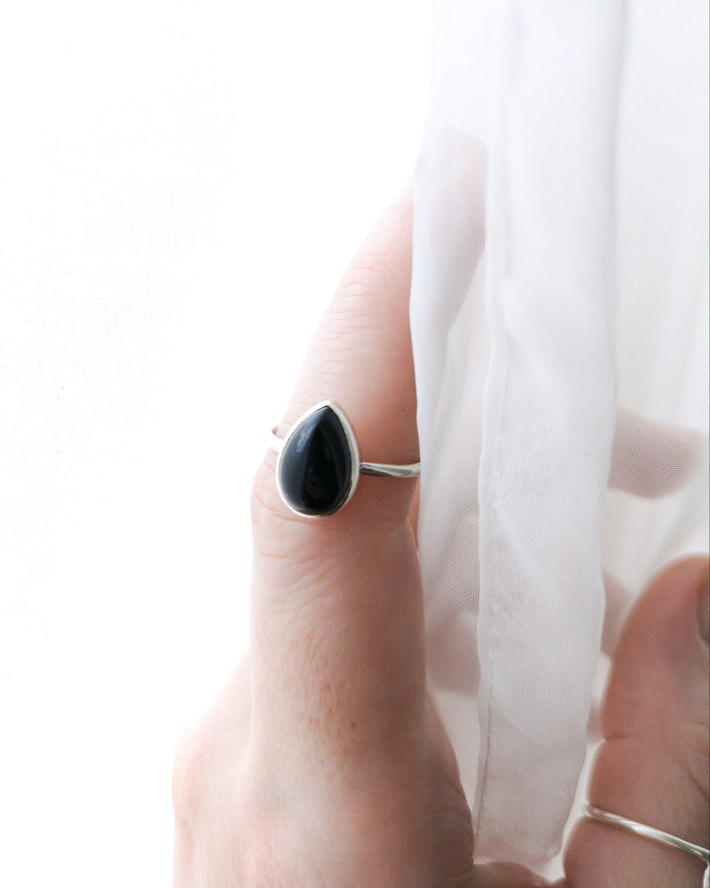 El anillo ESENCIAL de plata con una piedra de ónix negra en forma de lágrima, engarzada en un bisel simple, se encuentra en el dedo anular de una mano, cerca de una tela blanca y translúcida.
