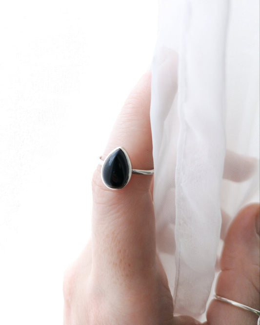 El anillo ESENCIAL de plata con una piedra de ónix negra en forma de lágrima, engarzada en un bisel simple, se encuentra en el dedo anular de una mano, cerca de una tela blanca y translúcida.