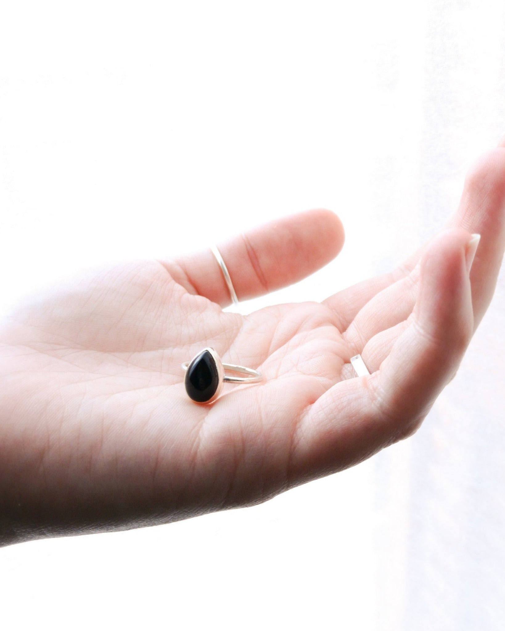 El anillo ESENCIAL de plata con una piedra de ónix negra en forma de lágrima, engarzada en un bisel simple, descansa sobre la palma abierta de una mano.