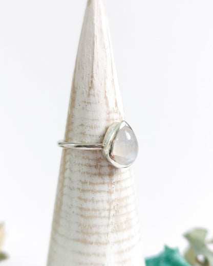 El anillo ESENCIAL de plata con una piedra lunar ovalada y translúcida, engarzada en un bisel simple, descansa sobre un expositor de anillo cónico de madera clara.