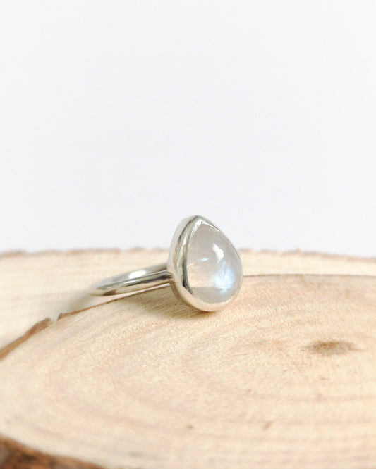 El anillo ESENCIAL de plata con una piedra lunar ovalada y translúcida, engarzada en un bisel simple, descansa sobre un trozo de madera con vetas visibles.
