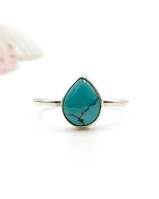 El anillo Sentir de la colección Esencial con una piedra de turquesa en forma de lágrima, engarzada en un bisel simple, sobre un fondo blanco con un detalle rosa difuminado en la esquina superior izquierda.