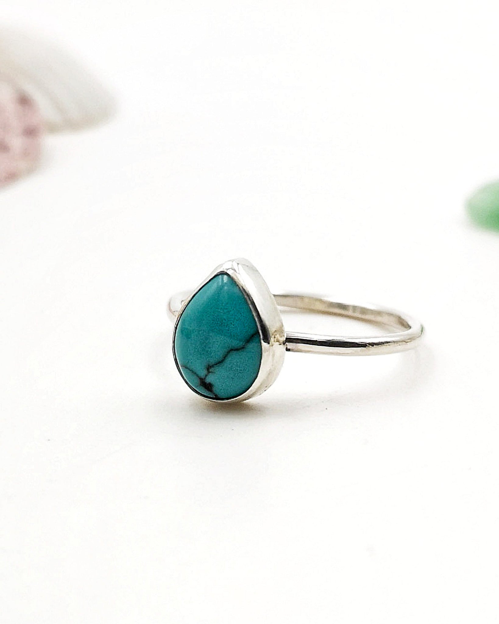El anillo Sentir de la colección Esencial con una piedra de turquesa en forma de lágrima, engarzada en un bisel simple, de medio perfil, sobre un fondo blanco con un detalle rosa difuminado en la esquina superior izquierda.
