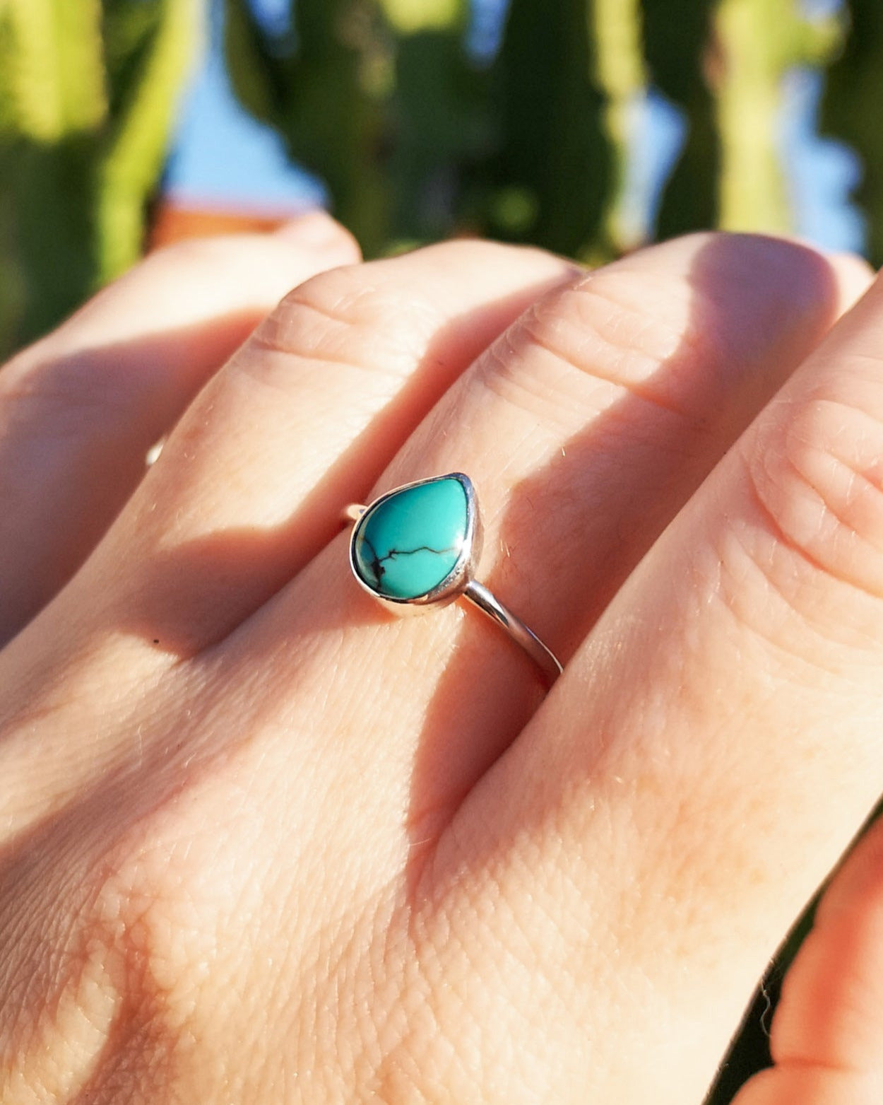 Mano usando en el dedo medio el anillo esencial sentir con piedra turquesa en forma de gota, de fondo el verde de un árbol.