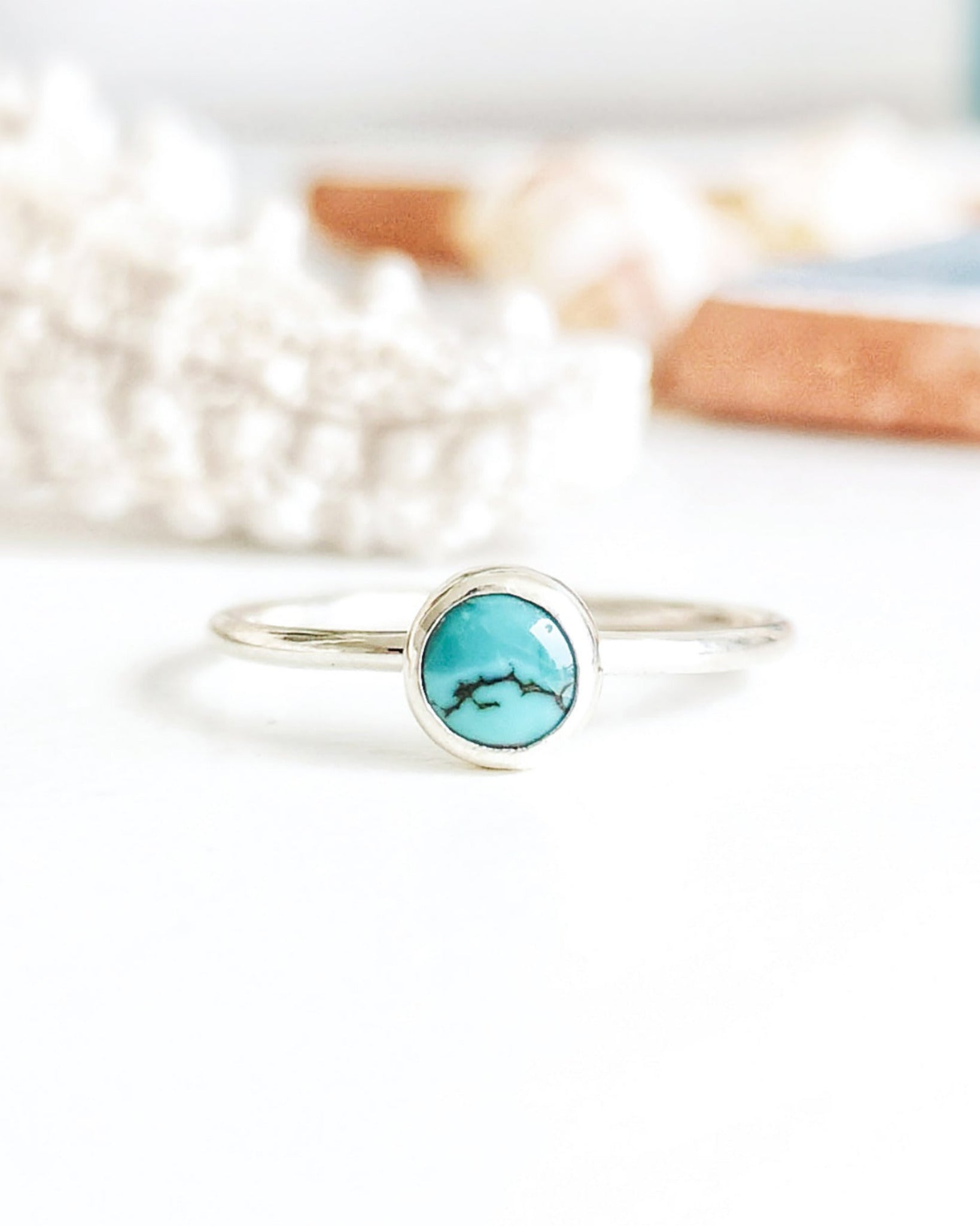 Anillo de plata con una piedra turquesa redonda en el centro, apoyado sobre una superficie blanca con objetos decorativos blancos y tonos tierra difuminados al fondo.