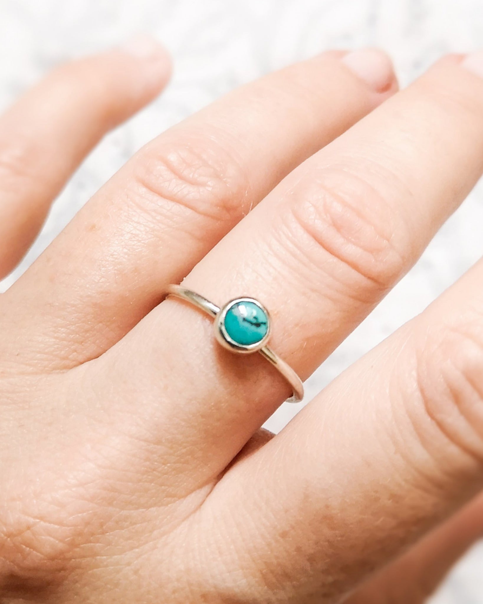 Anillo de plata con una piedra turquesa redonda en el centro, llevado en el dedo anular de una mano sobre un fondo claro y ligeramente texturizado.
