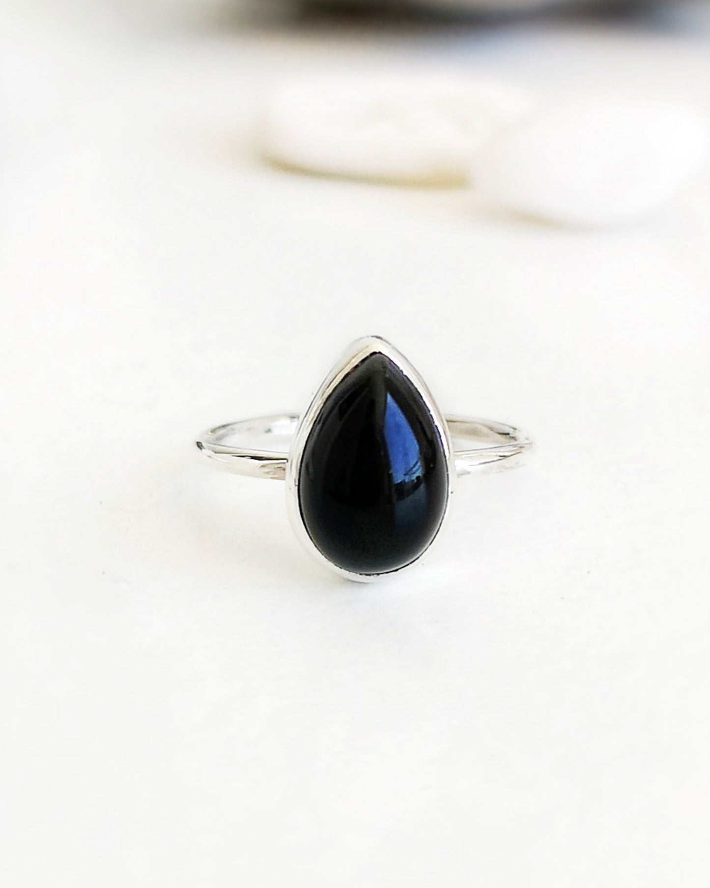 El anillo ESENCIAL de plata con una piedra de ónix negra en forma de lágrima, engarzada en un bisel simple, se encuentra en primer plano sobre una superficie blanca, con algunas piedras blancas y una pieza de madera desenfocadas al fondo.