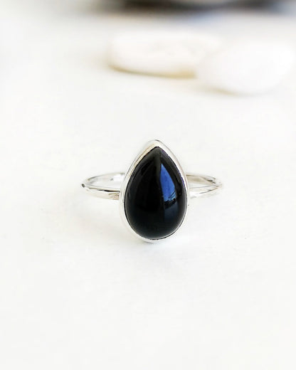 El anillo ESENCIAL de plata con una piedra de ónix negra en forma de lágrima, engarzada en un bisel simple, se encuentra en primer plano sobre una superficie blanca, con algunas piedras blancas y una pieza de madera desenfocadas al fondo.