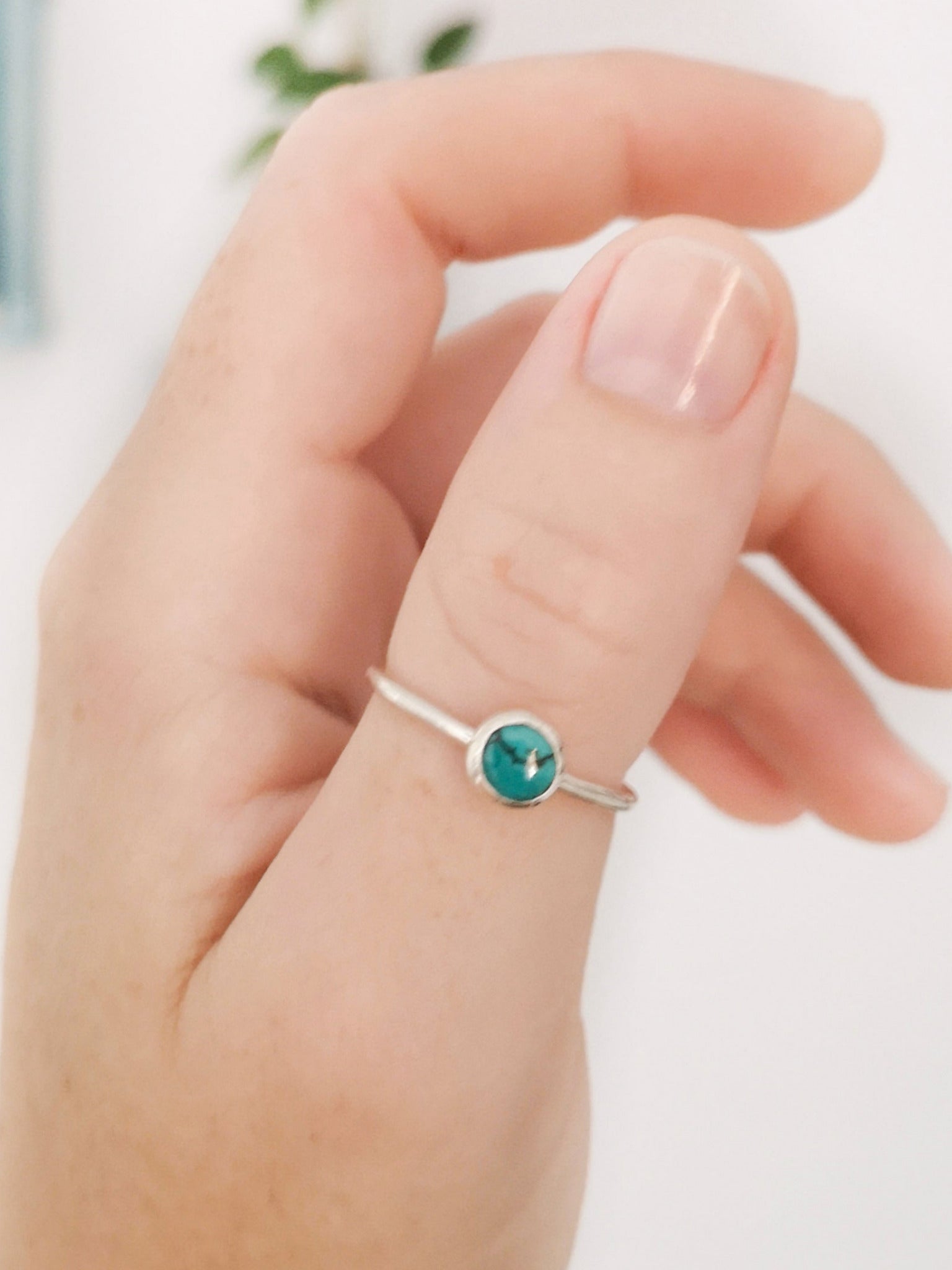 Anillo de plata con una piedra turquesa redonda en el centro, llevado en el dedo pulgar de una mano, con una planta y una estantería azul difuminadas al fondo.