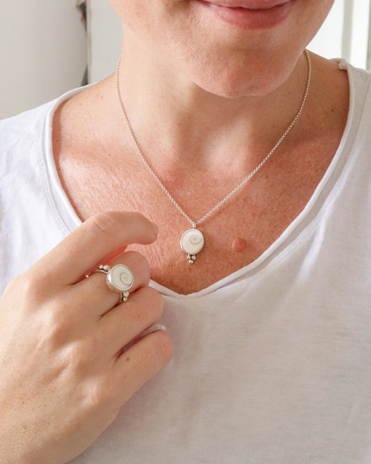Una persona lleva puesto un collar con un colgante redondo de plata y un anillo a juego, ambos con un diseño de espiral blanca en el centro, sobre una camiseta blanca.
