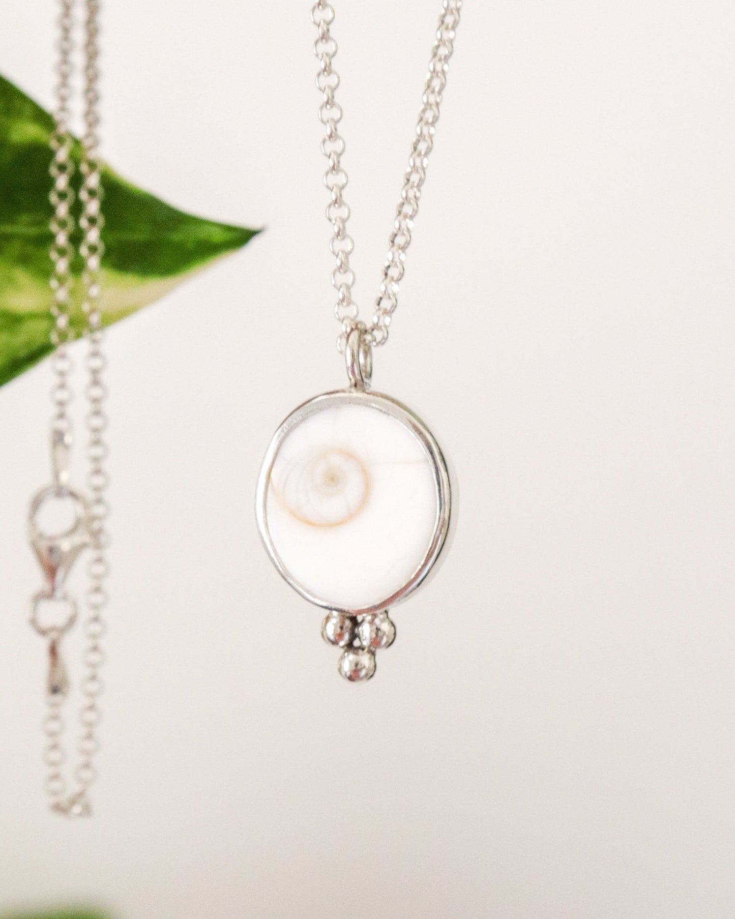 Un colgante redondo de plata con un diseño de espiral blanca en el centro y tres pequeñas esferas de plata colgando en la parte inferior, suspendido de una cadena de plata fina con un fondo difuminado.