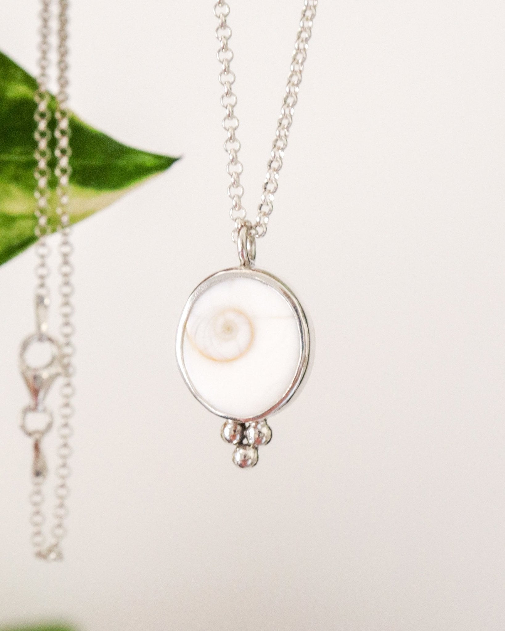 Un colgante redondo de plata con un diseño de espiral blanca en el centro y tres pequeñas esferas de plata colgando en la parte inferior, suspendido de una cadena de plata fina con un fondo difuminado.