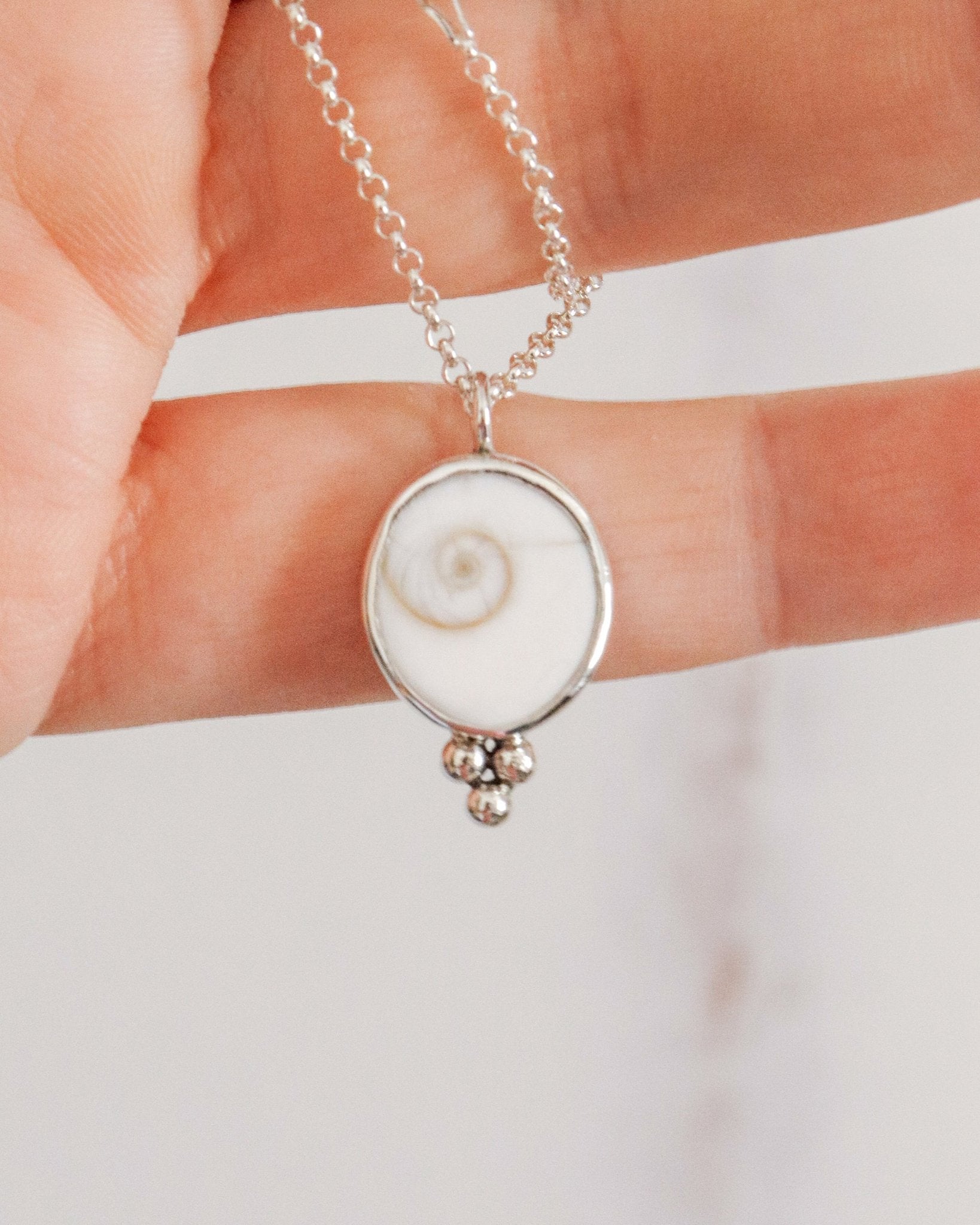 Una mano sostiene un colgante redondo de plata con un diseño de espiral blanca en el centro y tres pequeñas esferas de plata en la parte inferior, colgado de una cadena fina de plata.