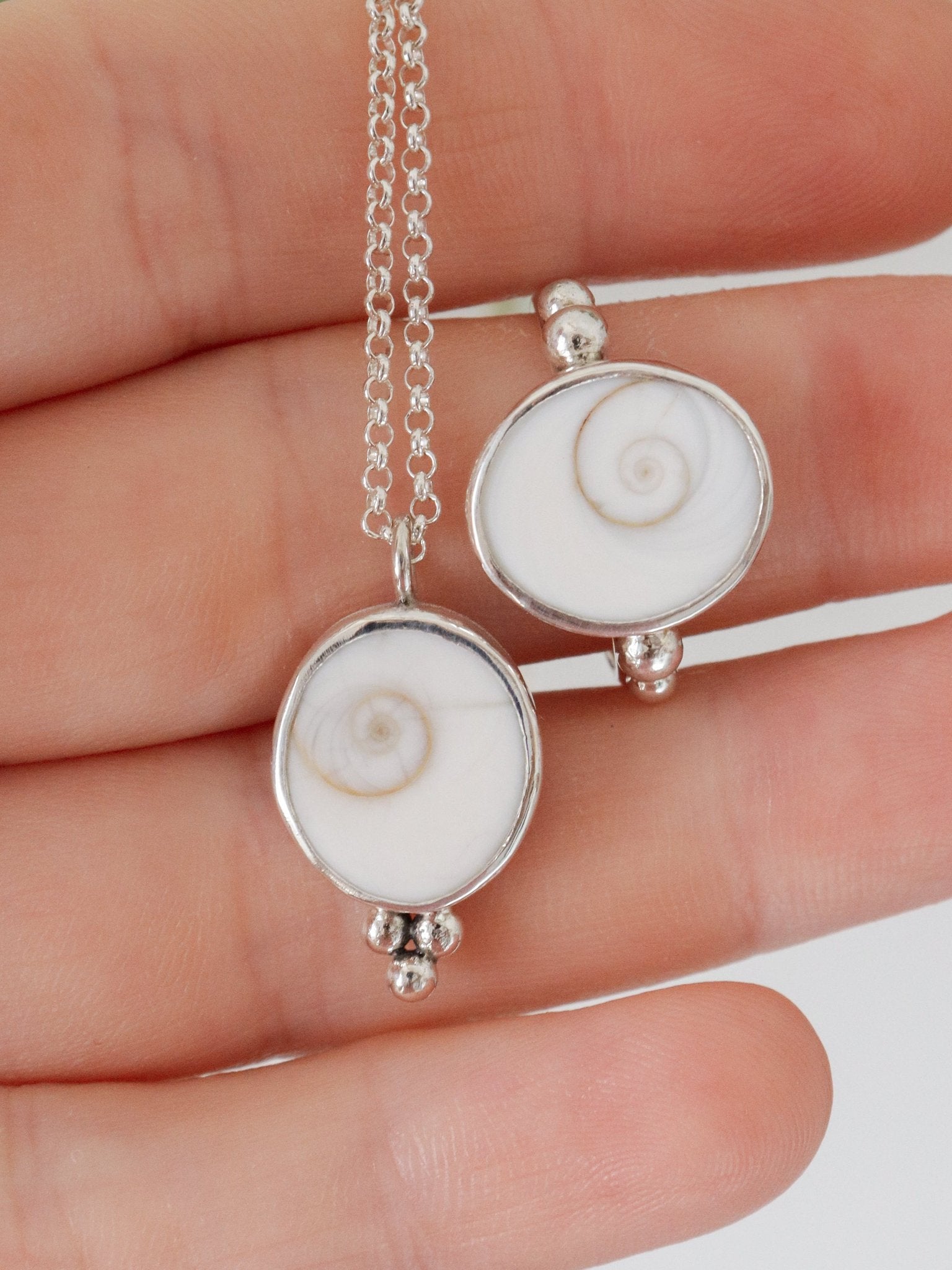 Colgante y anillo de plata, cada uno con un opérculo de caracol blanco con un diseño de espiral y tres pequeñas esferas de plata colgando, se muestran sobre la palma de una mano. El colgante está unido a una cadena de plata de ley.