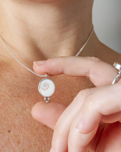 Una persona lleva puesto un collar con un colgante redondo de plata que contiene un opérculo blanco con espiral, con la mano tocando suavemente el colgante.