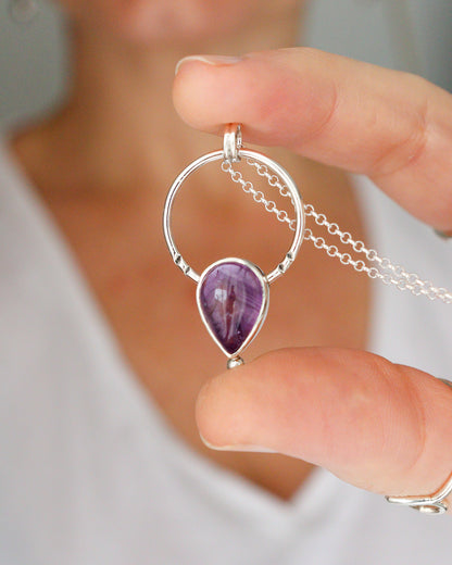 Una persona sostiene el colgante ARCO, una joya de plata con un diseño circular que enmarca una piedra preciosa de amatista en forma de lágrima, con una cadena fina de plata visible.