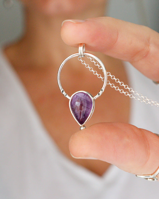 Una persona sostiene el colgante ARCO, una joya de plata con un diseño circular que enmarca una piedra preciosa de amatista en forma de lágrima, con una cadena fina de plata visible.