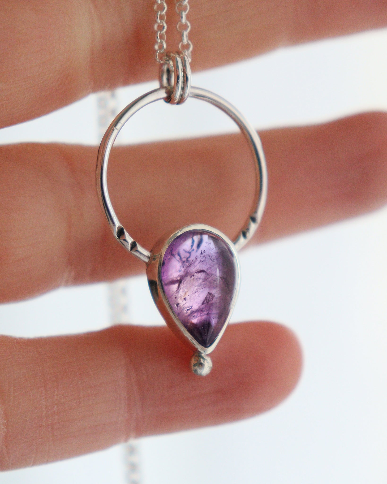 Una vista de cerca del colgante ARCO de plata, con un arco circular que enmarca una piedra de amatista en forma de lágrima, sostenido por una mano y con una cadena de plata fina que se extiende hacia la parte superior.