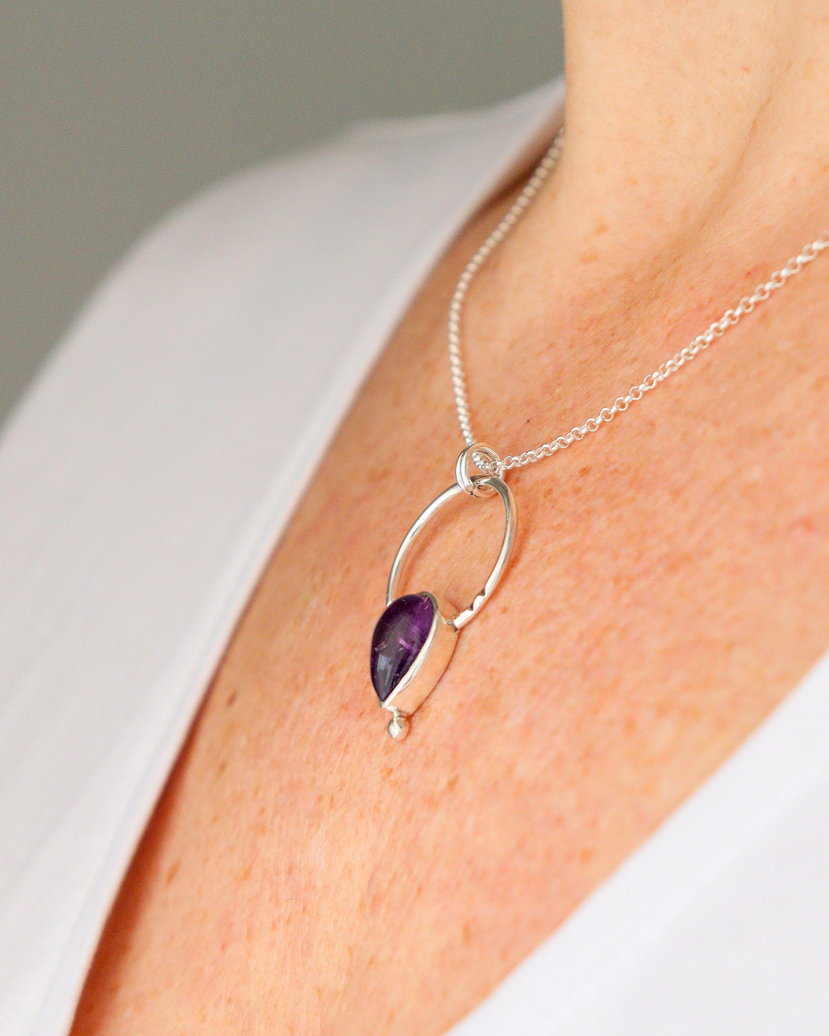 Una persona lleva puesto un collar con el colgante ARCO de plata, con un diseño curvo que abraza una piedra preciosa de amatista en forma de lágrima, sobre una camiseta blanca.