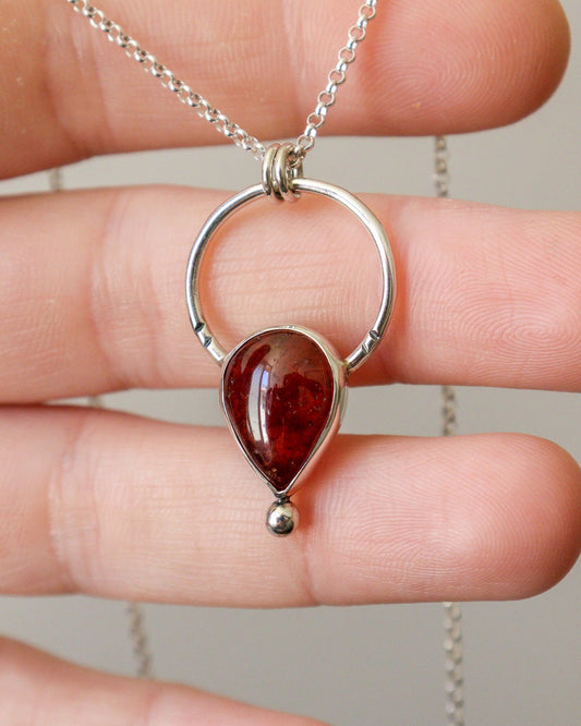 Una mano sostiene el colgante ARCO de plata con un círculo y una piedra preciosa de granate rojo intenso en forma de lágrima, colgado de una fina cadena de plata.