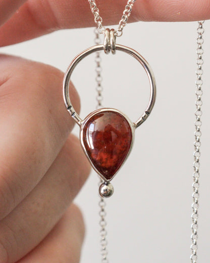 Un primer plano del colgante ARCO de plata con una piedra preciosa de granate rojo intenso en forma de lágrima, con una cadena de plata fina desenfocada en el fondo.