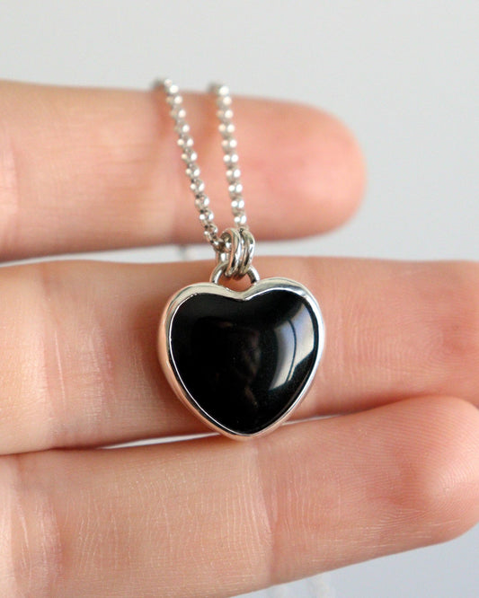 Una mano sostiene un collar con un colgante en forma de corazón de ónix negro brillante engarzado en plata, colgado de una cadena de plata.