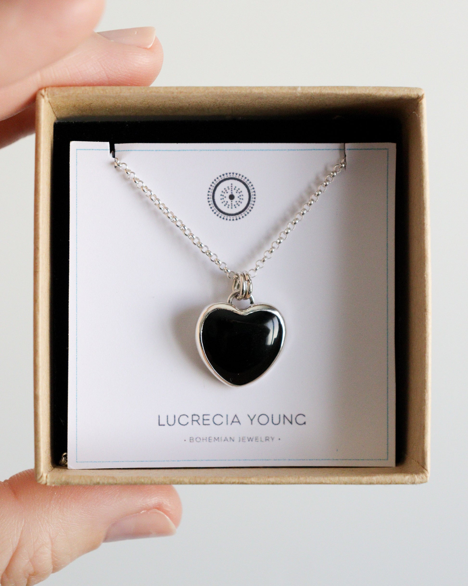 Una mano sostiene una pequeña caja de cartón abierta con un collar de plata y un colgante de ónix negro en forma de corazón, sobre un soporte de cartulina blanca con el logotipo "LUCRECIA YOUNG - BOHEMIAN JEWELRY".