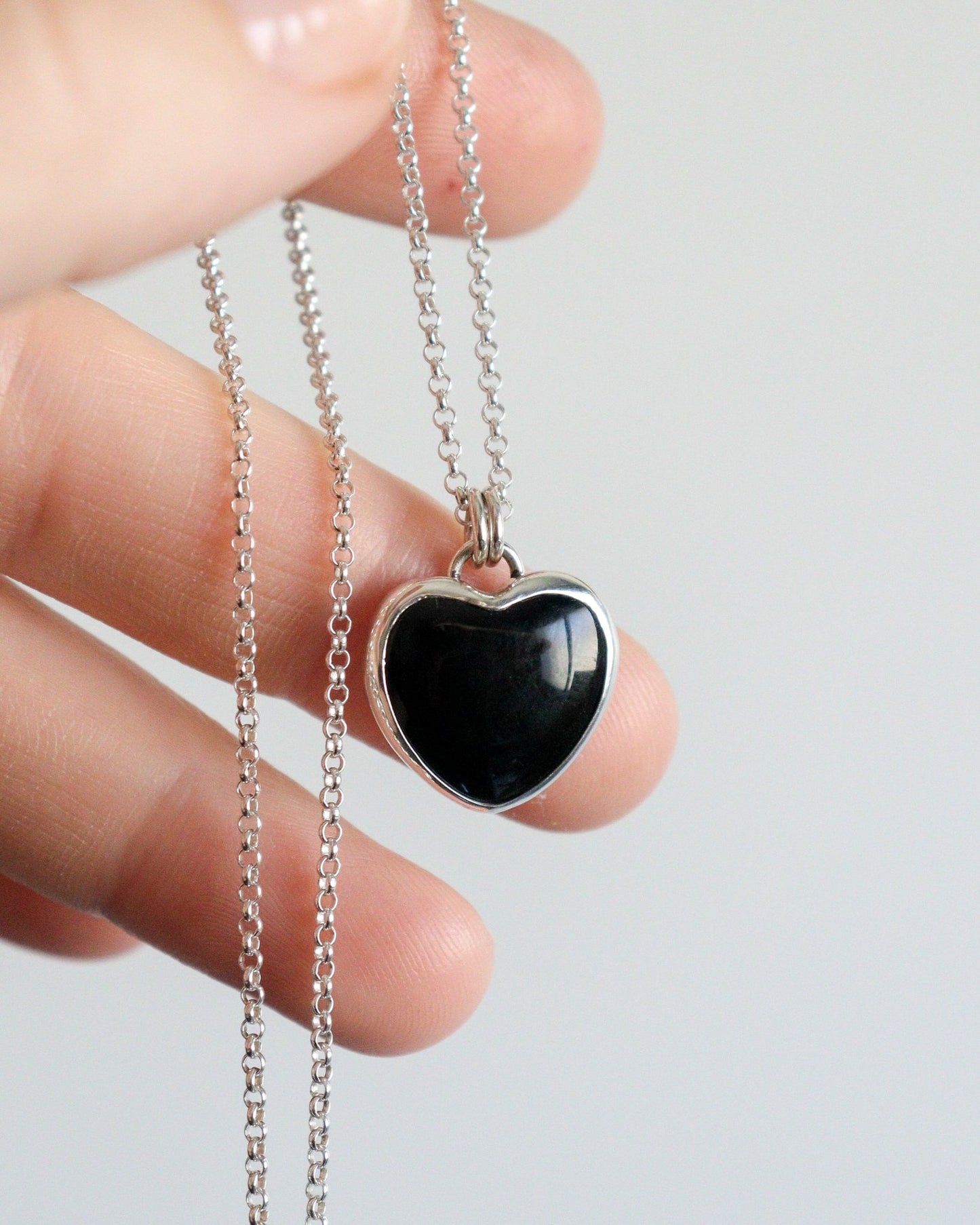 Una mano sostiene un collar con un colgante de ónix negro en forma de corazón, engarzado en plata, y su cadena de plata fina.