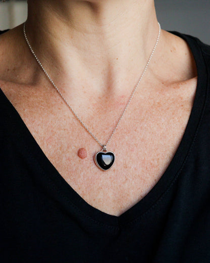 Una persona lleva puesto un collar con un colgante de ónix negro en forma de corazón, engarzado en plata, sobre una camiseta oscura.
