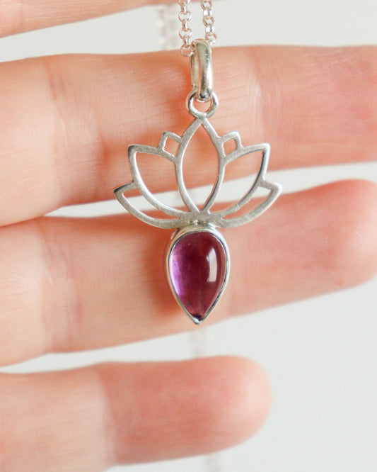 Un colgante de plata con diseño de flor de loto y una amatista en forma de lágrima, sostenido por una mano.