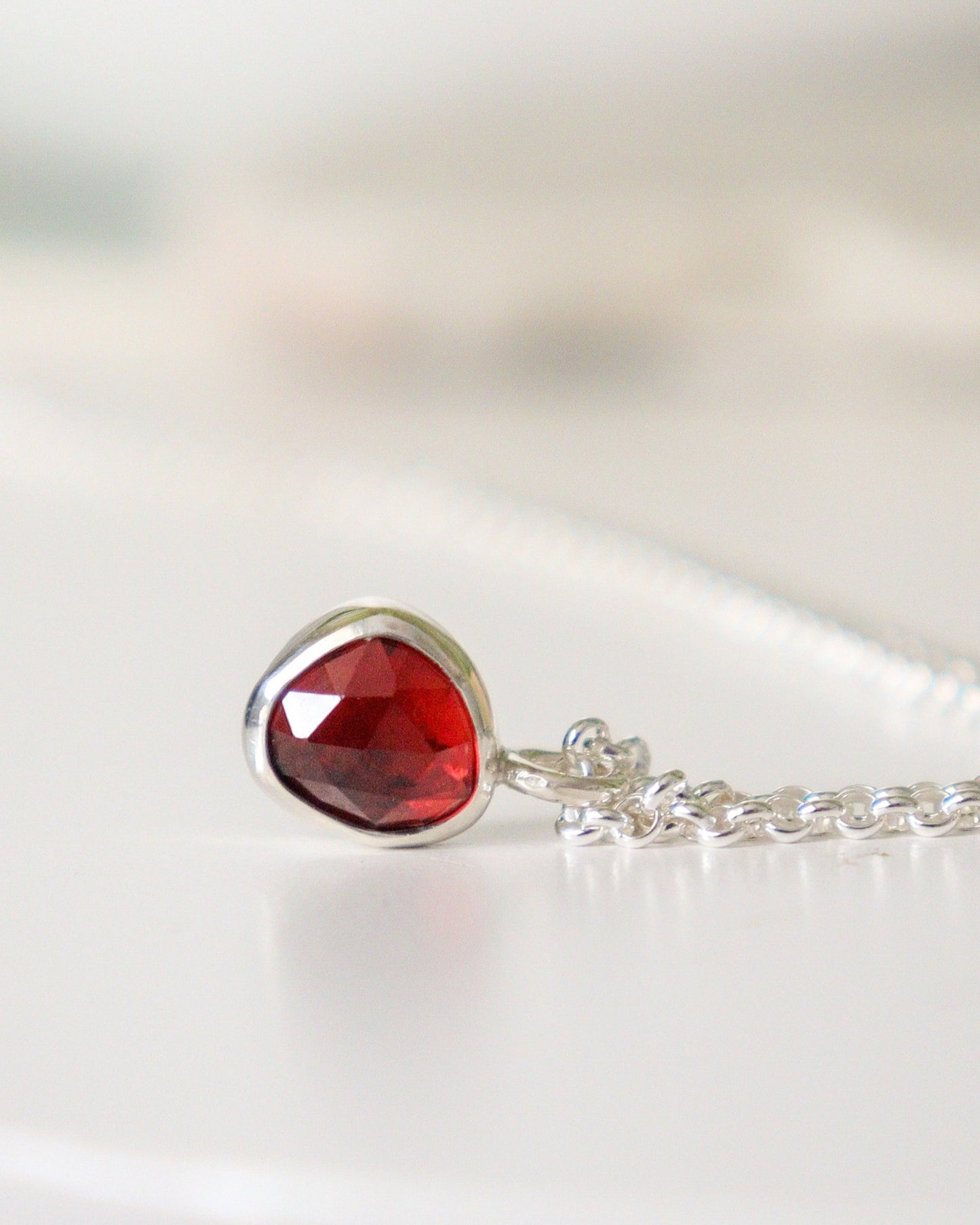 Colgante de mini granate rojo de talla rosa reposa con su cadena sobre una superficie blanca.