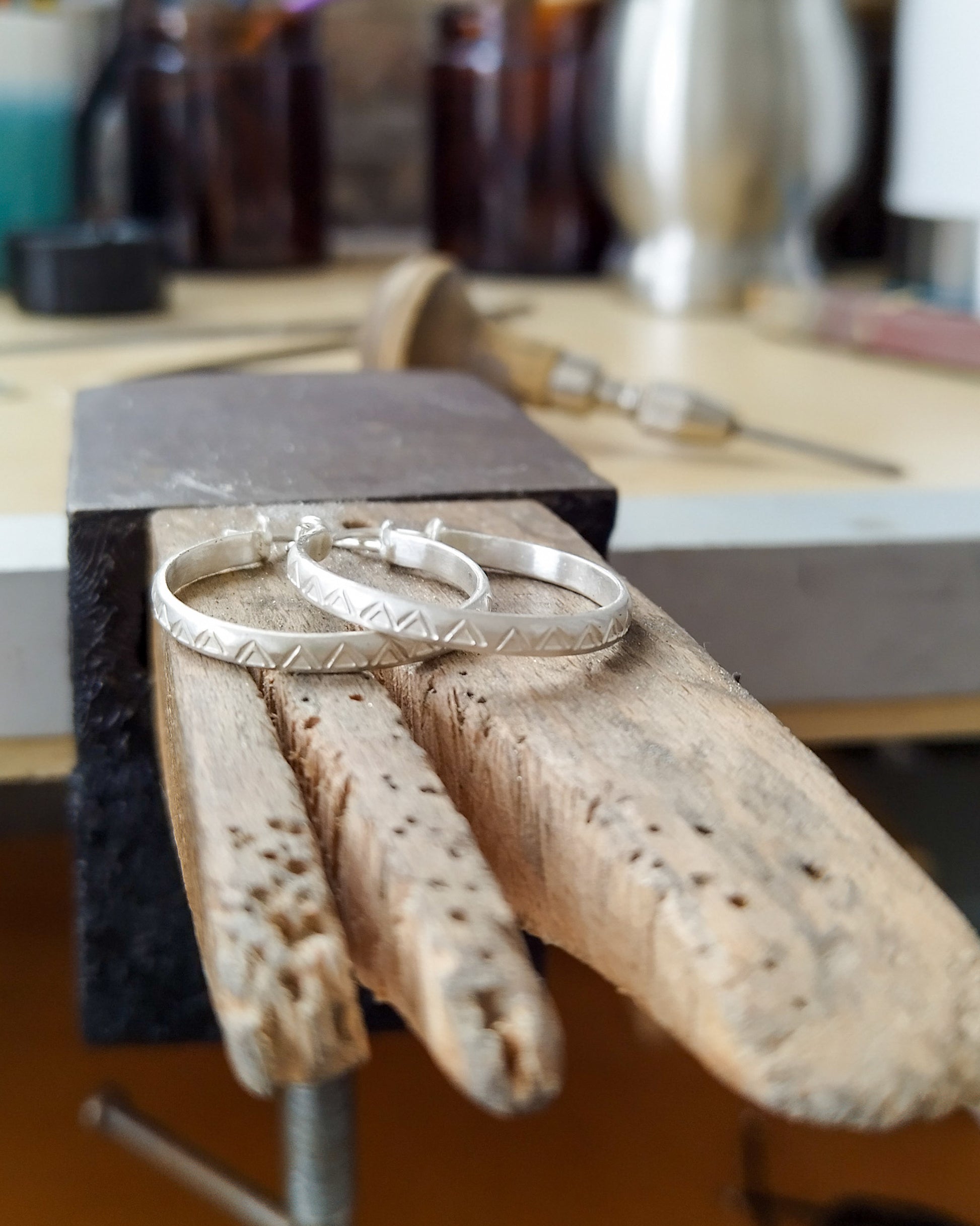 Un par de pendientes de aro de plata con un patrón de triángulos grabados (pendientes de aro TRILO) descansan sobre un trozo de madera de banco de joyero, que tiene varias muescas y agujeros. Al fondo, se pueden ver herramientas de joyería y botellas en un entorno de taller desenfocado.