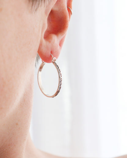 Primer plano de una oreja con un pendiente de aro plateado colgado del lóbulo. El aro tiene un patrón triangular grabado alrededor de su borde inferior (pendientes de aro TRILO). Se puede apreciar un segundo pendiente de aro más pequeño en la parte superior de la oreja. El fondo es claro y ligeramente borroso.