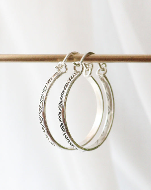 Dos pendientes de aro de plata idénticos, con un patrón geométrico de triángulos grabado (pendientes TRILO), cuelgan de una barra de metal horizontal de color dorado o bronce. Los pendientes están bien enfocados y el fondo es liso y ligeramente desenfocado.