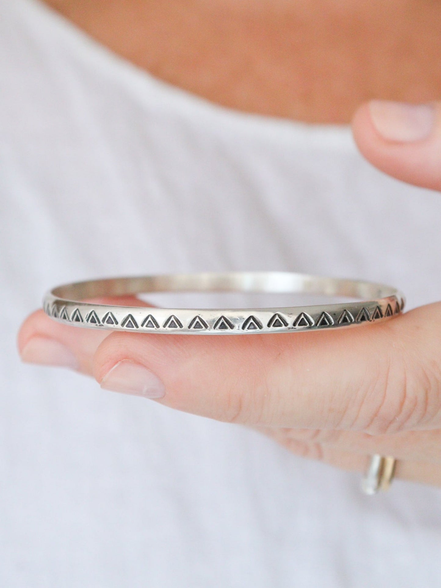 Primer plano de la pulsera TRILO de plata de ley con un diseño minimalista y tribal. El brazalete redondo tiene un patrón de triángulos estampados que se extiende por toda la pieza. La pulsera es sostenida por una mano sobre una blusa blanca.