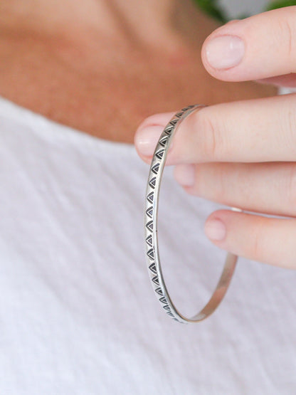 Primer plano de la pulsera TRILO de plata de ley minimalista con un detalle tribal. La pulsera es un brazalete redondo con un patrón de triángulos estampados, sostenida por una mano.