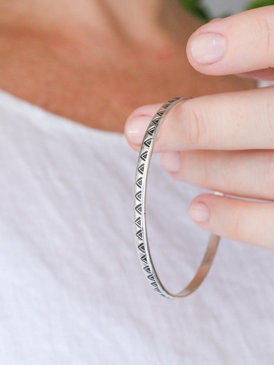 Primer plano de la pulsera TRILO de plata de ley minimalista con un detalle tribal. La pulsera es un brazalete redondo con un patrón de triángulos estampados, sostenida por una mano.