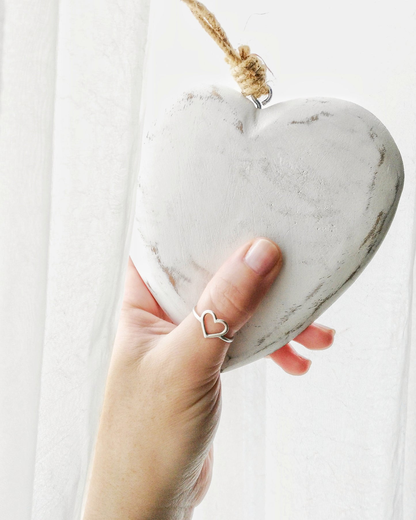 Imagen en primer plano de mano de perfil sosteniendo un corazón de madera color blanco. La mano lleva el anillo CORAZÓN en el dedo pulgar. Fondo blanco.